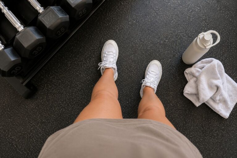 Top-down view of legs in white gym shoes standing next to dumbbells, a water bottle, and a towel on a gym floor.